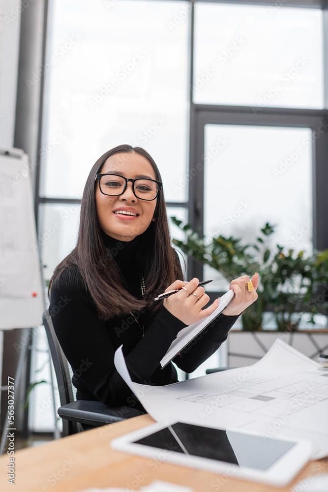 Asian designer smiling at camera and holding notebook near blueprint and digital tablet. Asian designer smiling at camera and holding notebook near blueprint and digital tablet.