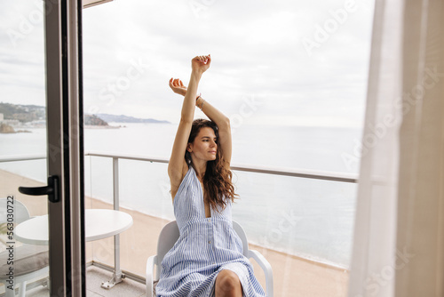 Free Download Pretty young caucasian girl dressed striped cocktail dress relaxing on hotel balcony with beach in background. Brown hair woman sitting on chair of residence. Concept happy weekend, tourist. - GFXLoad