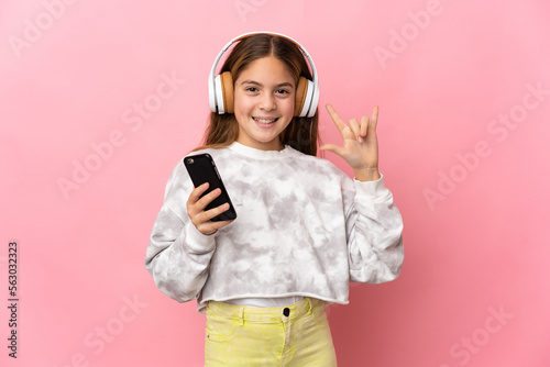 Child over isolated pink background listening music with a mobile making rock gesture