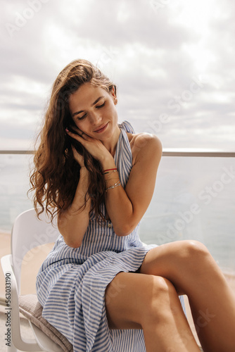 Tender young caucasian girl sits on chair spending leisure time at balcony. Brunette is wearing blue dress outside. Relax, vacation concept.