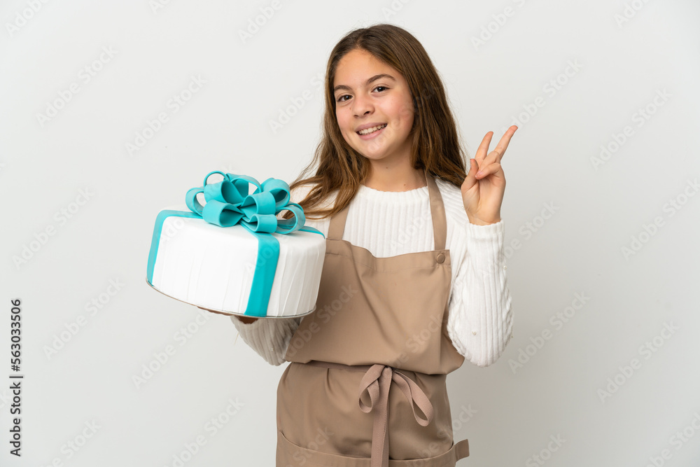 Little girl holding a big cake over isolated white background smiling and showing victory sign Little girl holding a big cake over isolated white background smiling and showing victory sign