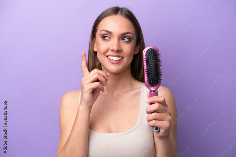 Young caucasian woman holding hairbrush isolated on purple background intending to realizes the solution while lifting a finger up Young caucasian woman holding hairbrush isolated on purple background intending to realizes the solution while lifting a finger up