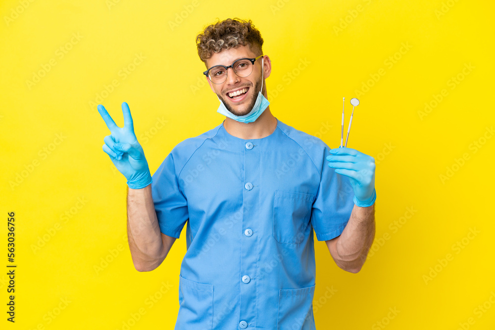 Dentist blonde man holding tools isolated on background smiling and showing victory sign