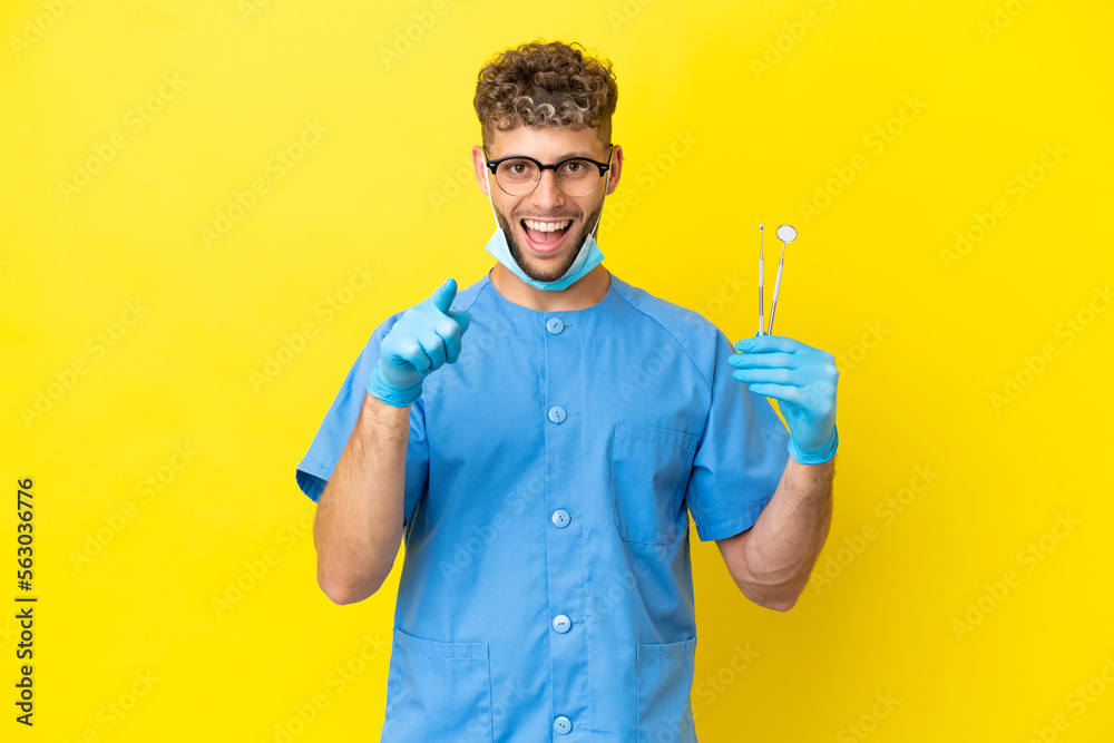 Dentist blonde man holding tools isolated on background surprised and pointing front Dentist blonde man holding tools isolated on background surprised and pointing front
