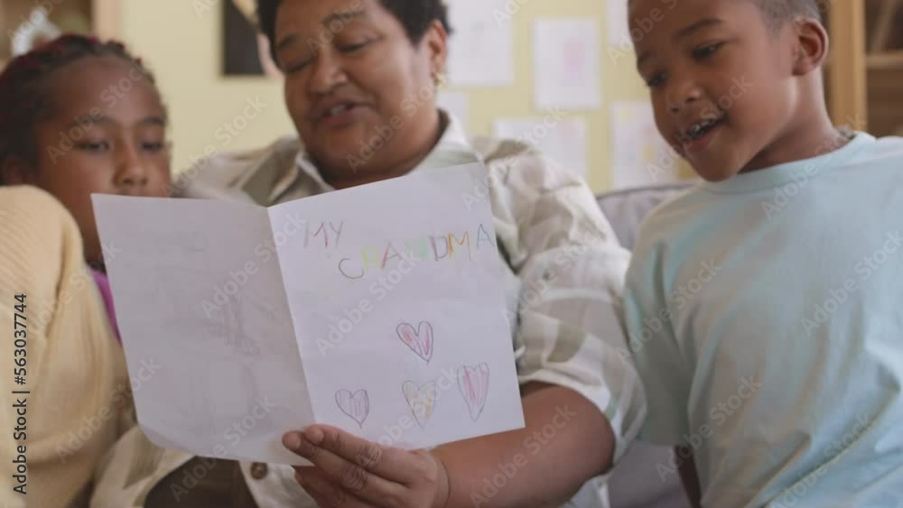 Mature Black woman reading postcard made for her by grandchildren sitting together on couch in living room Mature Black woman reading postcard made for her by grandchildren sitting together on couch in living room