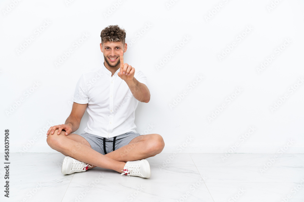 Young blonde man sitting on the floor isolated on white background showing and lifting a finger Young blonde man sitting on the floor isolated on white background showing and lifting a finger