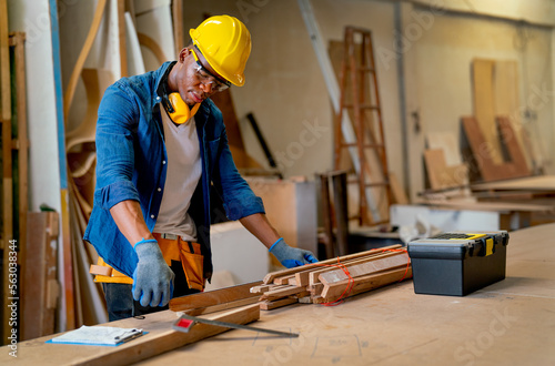 African American carpenter man use measure tape to check and work with timber in factory workplace and tools box is place on table near working area. photo