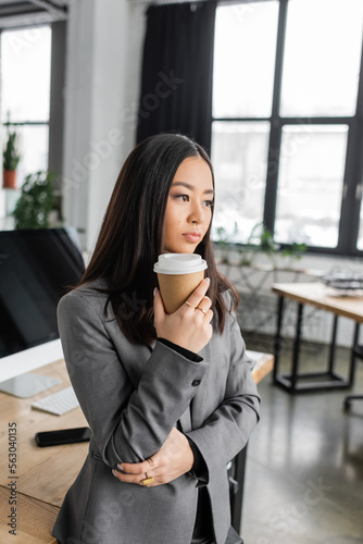 Brunette asian interior designer holding paper cup while thinking in office.