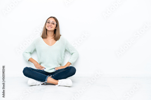 Young caucasian woman sitting on the floor isolated on white background posing with arms at hip and smiling