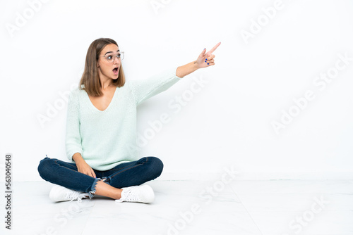 Young caucasian woman sitting on the floor isolated on white background pointing away
