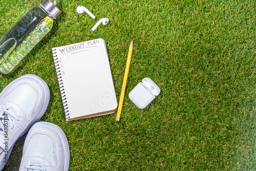 Spring outdoor sport and fitness flat lay. Sport shoes, dumbbells, blank notebook for workout plan, earphones, water bottle on sunny green grass background, top view copy space