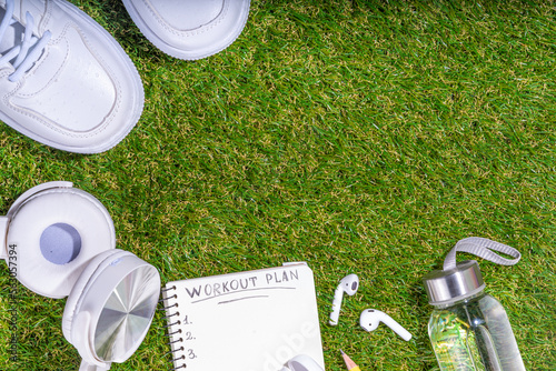 Spring outdoor sport and fitness flat lay. Sport shoes, dumbbells, blank notebook for workout plan, earphones, water bottle on sunny green grass background, top view copy space