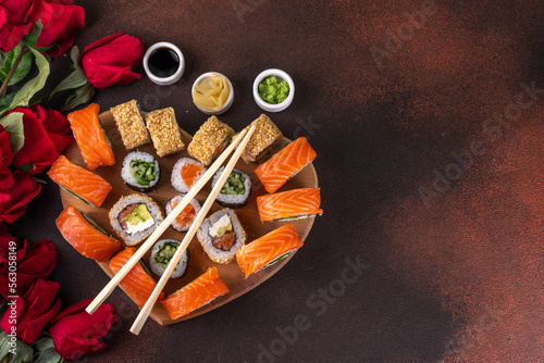 Heart shaped Valentine day sushi set. Classic sushi rolls, philadelphia, maki set for two, with two pairs of chopsticks for Valentine's dating dinner, with rose flowers bouquet on dark background