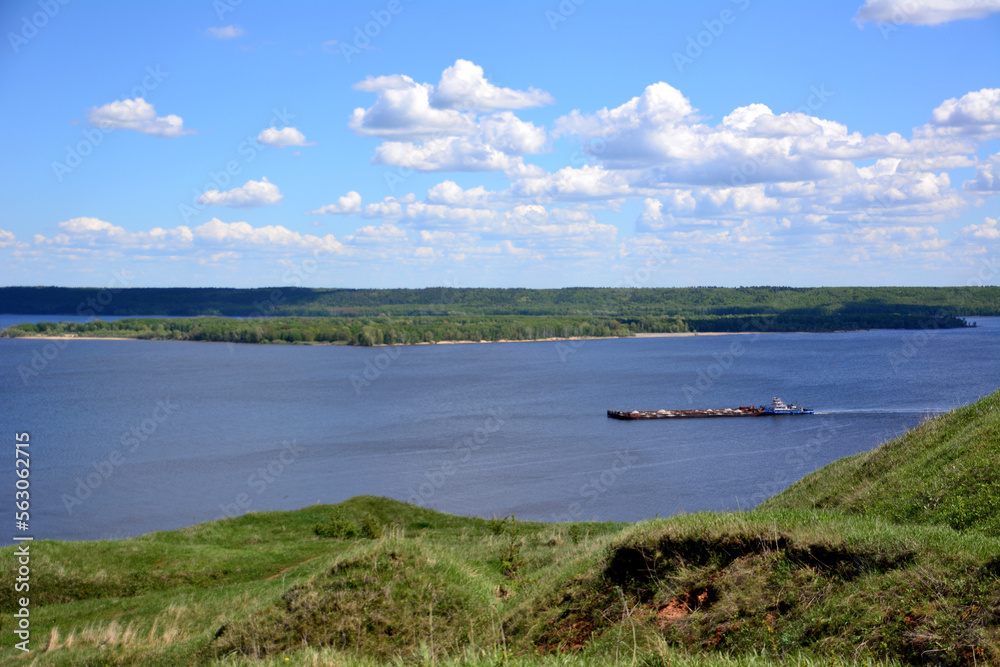 barge on the Volga river swimming forward with green hills and blue sky and clouds