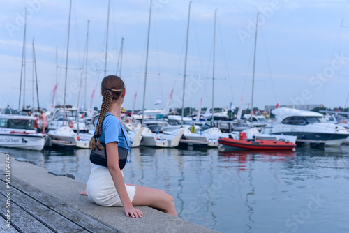 The girl sits on the pier in the port and looks at the boats and yachts.