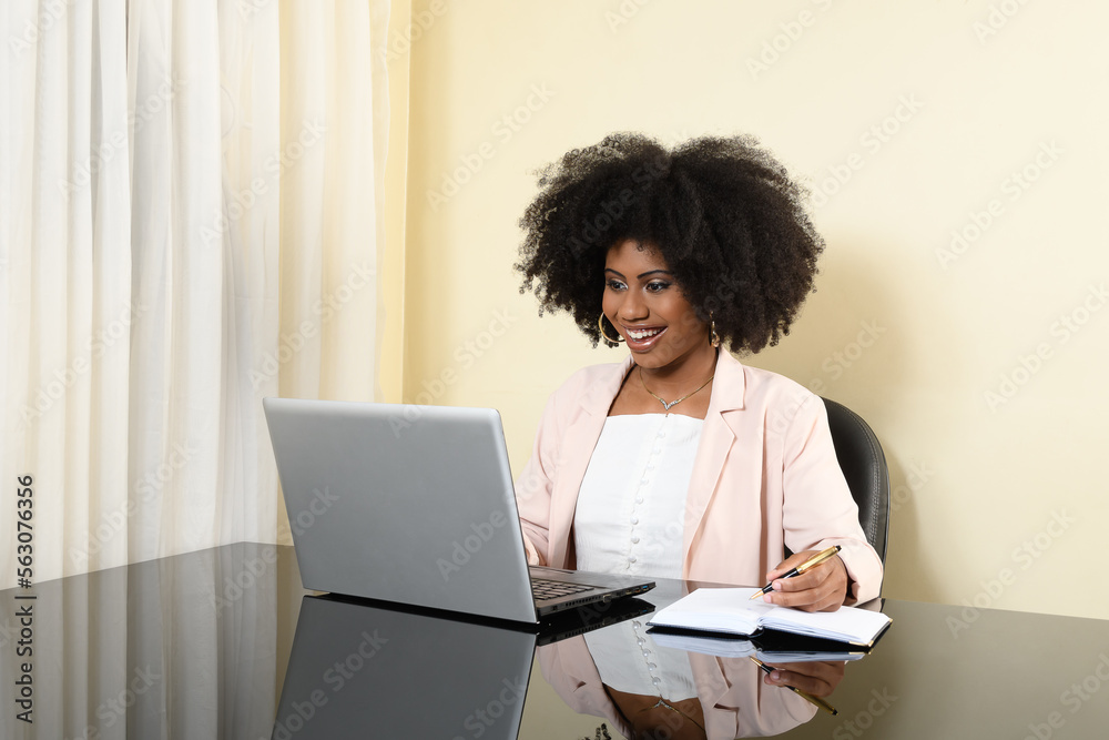 young woman sitting at her desk typing on her laptop camputer keyboard looks at screen and smiles, black woman