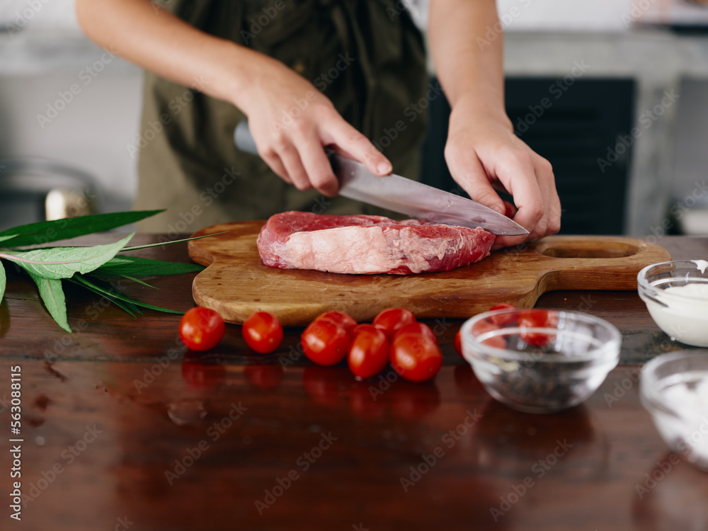 Woman with knife in hand cutting fresh steak meat for roasting in kitchen with salt pepper and other spices on table, red cherry tomatoes and herbs, preparing dinner.