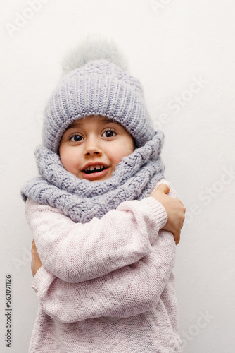 child girl in a gray hat and scarf hugs herself on a white background and smiles, grimace, childhood, winter season