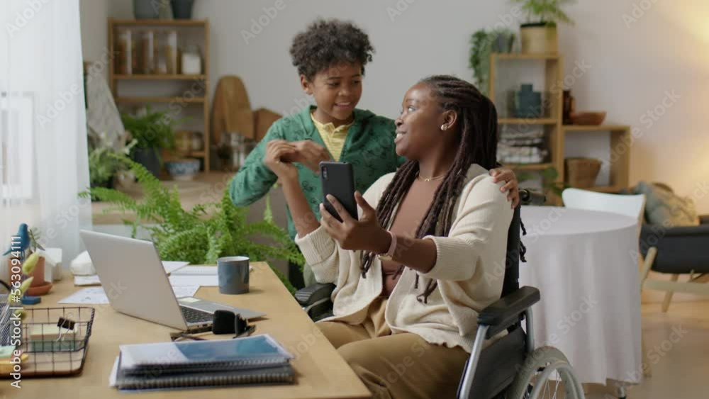 Medium long shot of African American woman in wheelchair smiling and posing for smartphone camera with little son while taking selfie together at home Medium long shot of African American woman in wheelchair smiling and posing for smartphone camera with little son while taking selfie together at home