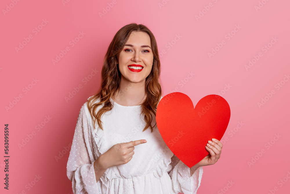 Valentine's Day concept. Caucasian smiling girl holding valentine card pointing finger at big paper heart isolated on pink background. Valentine's Day concept. Caucasian smiling girl holding valentine card pointing finger at big paper heart isolated on pink background.