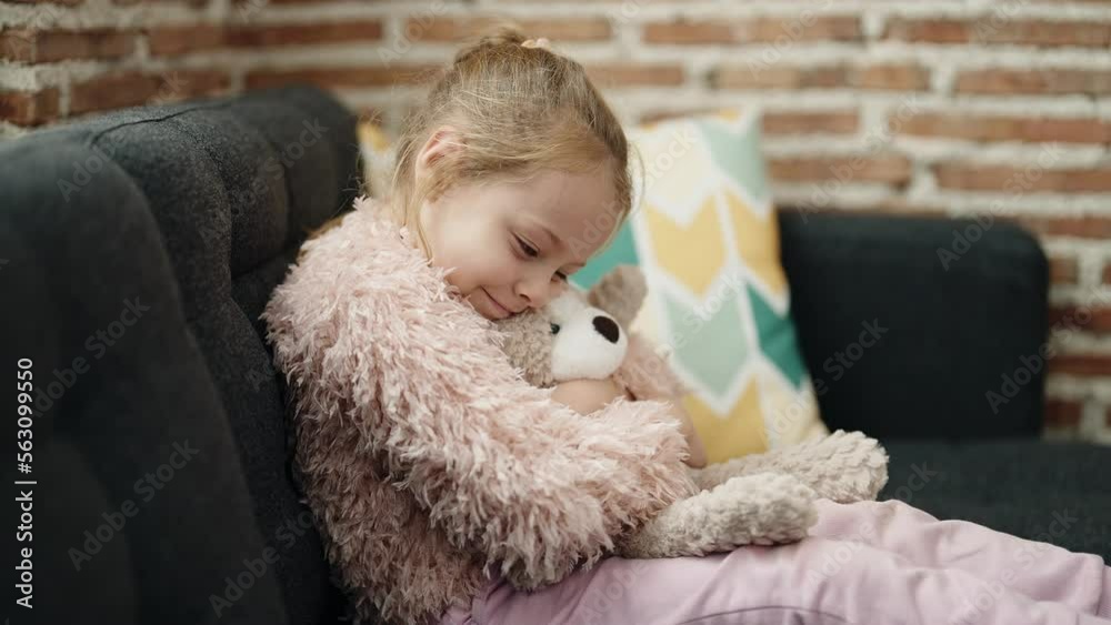 Adorable blonde girl hugging teddy bear sitting on sofa at home Adorable blonde girl hugging teddy bear sitting on sofa at home