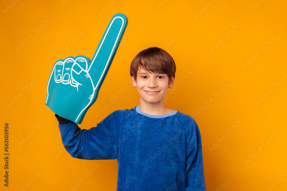 Handsome teen boy wearing blue foam fan finger glove smiles at the camera over yellow background. Handsome teen boy wearing blue foam fan finger glove smiles at the camera over yellow background.