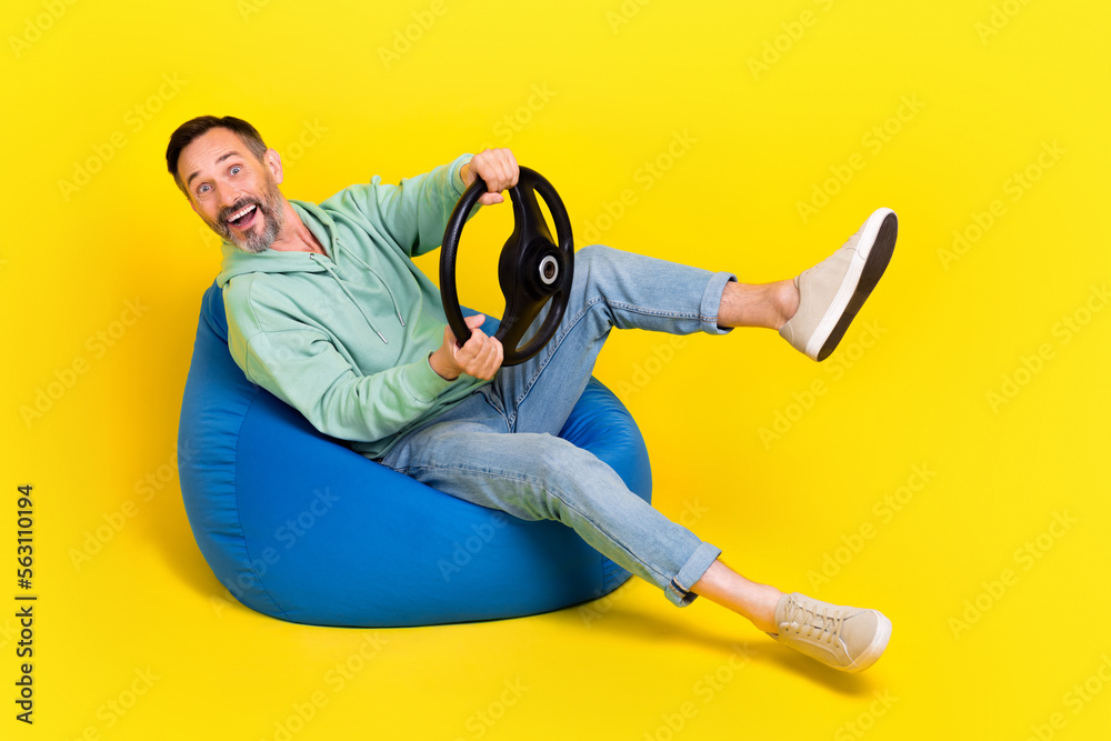 Full length photo of funny excited man wear green sweatshirt driving auto having fun sitting beanbag isolated yellow color background Full length photo of funny excited man wear green sweatshirt driving auto having fun sitting beanbag isolated yellow color background