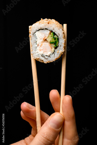 Close-up view of piece of roll california with snow crab meat, cream cheese, cucumber garnished sesame seeds in bamboo chopsticks in hand
