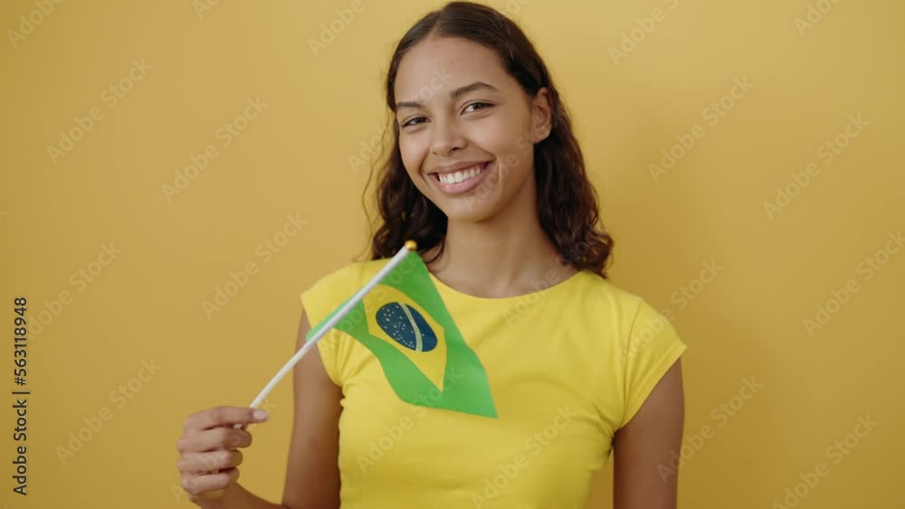 Young african american woman smiling confident holding brazil flag over isolated yellow background Young african american woman smiling confident holding brazil flag over isolated yellow background