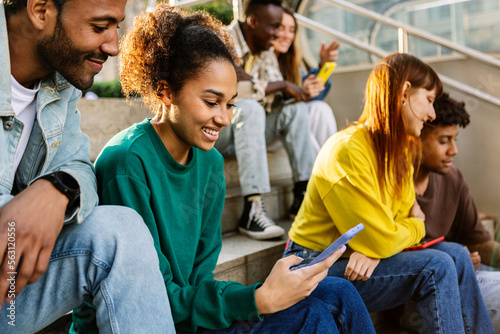 Young group of diverse student friends using mobile phone outdoor. Millennial generation people having fun watching social media content on smartphone app. Youth technology lifestyle concept