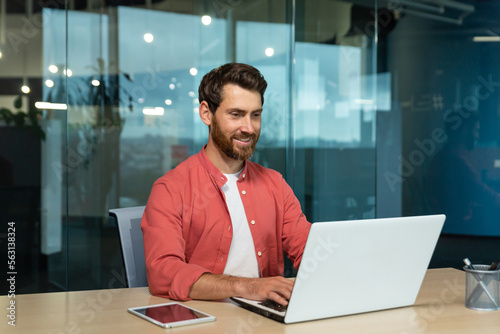 Successful businessman in red shirt happily working with laptop inside office, mature man with beard at workplace typing on keyboard smiling satisfied with work results and achievement.