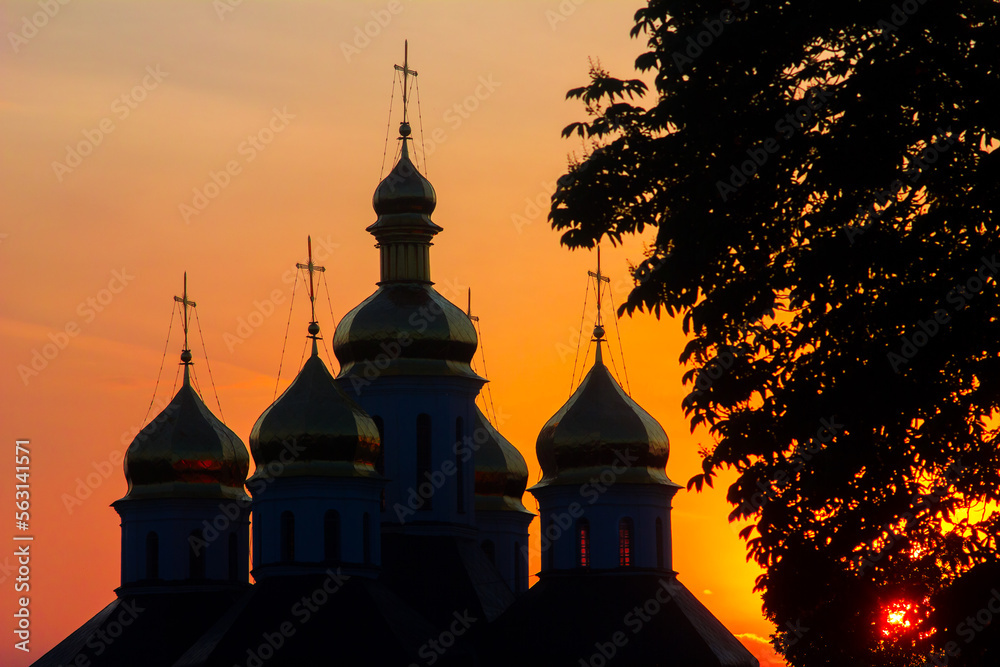 silhouette of an orthodox church or cathedral with domes and crosses against the background of a yellow-red fiery sunset and sunlight passing through the foliage of a tree in Ukraine in Europe