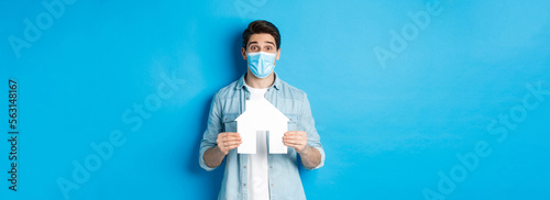 Concept of coronavirus, quarantine and social distancing. Young man searching apartment, showing house paper model, wearing medical mask, renting or buying propery, blue background photo