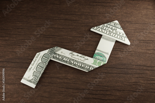 US 100 dollar bill folded into an up arrow on wooden table. Illustration of the concept of inflation, profit, earning and investment