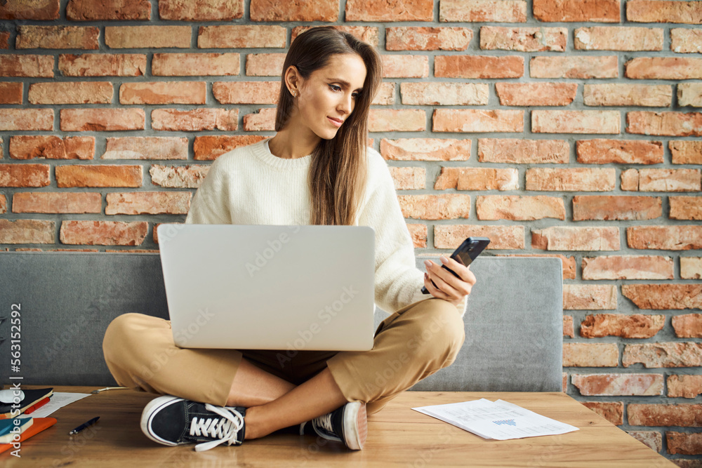 Casual businesswoman looking at phone while sitting with laptop on desk in office. Female entrepreneur doing mobile payment Casual businesswoman looking at phone while sitting with laptop on desk in office. Female entrepreneur doing mobile payment