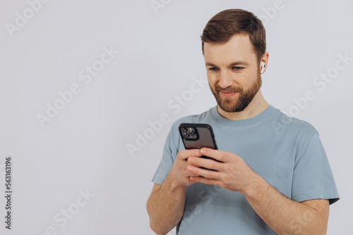 Portrait of a modern millennial man with a beard holding a smartphone and listening to something in wireless headphones on a white background with copy space © anatoliycherkas