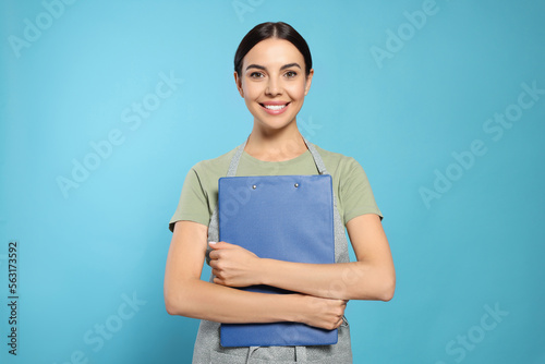 Young woman in grey apron with clipboard on light blue background