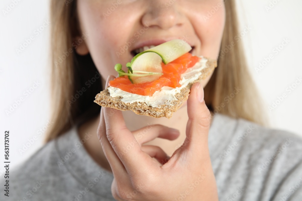 Woman eating fresh rye crispbread with cream cheese, salmon and cucumber on white background, closeup Woman eating fresh rye crispbread with cream cheese, salmon and cucumber on white background, closeup