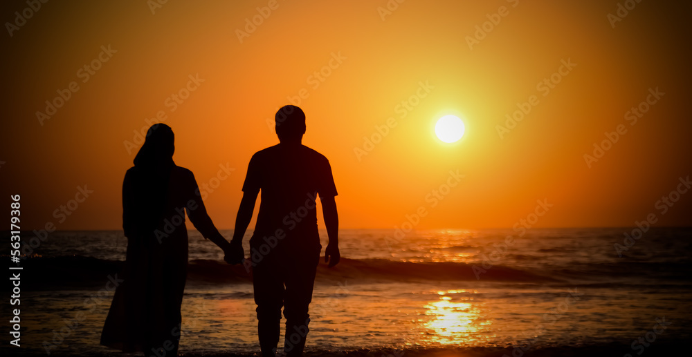 Couple in love watching sunset together on beach travel summer holidays. Silhouette of two young people (guy and girl) holding hands looking out to the sea at sunset.