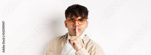 Close-up of grumpy young guy in glasses frowning and shushing, make taboo gesture with finger shuting lips, tell to be quiet, standing angry on white background photo