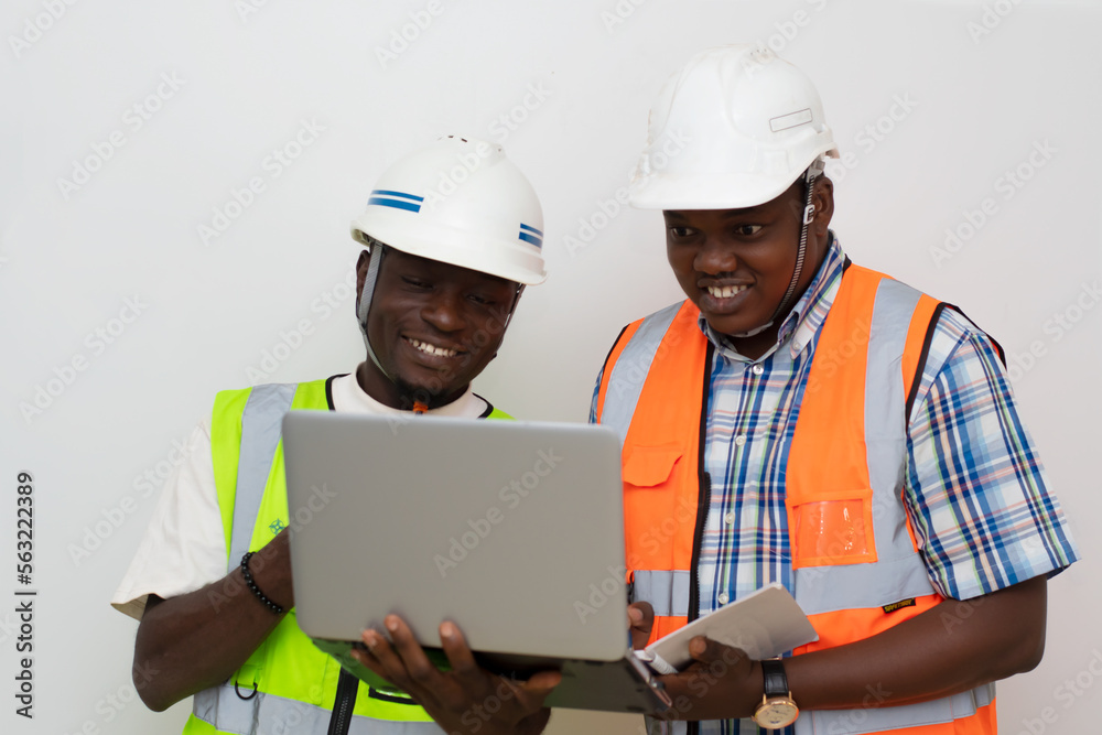Portrait of two african industrial builders standing at building site and looking at laptop display, smiling Portrait of two african industrial builders standing at building site and looking at laptop display, smiling