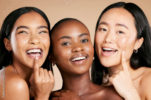 Skincare, beauty and multicultural women smile and point at skin on studio background. Health, wellness and luxury cosmetics, healthy care and happy, beautiful and friendly people with natural makeup