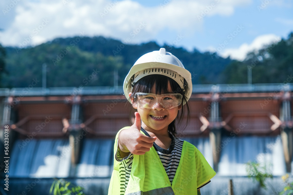 Portrait of a little girl engineer wearing a green vest and white helmet smiling happily on the background of the dam. Concepts of environmental engineering, renewable energy and love of nature.