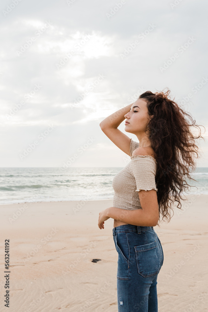 Side view of young woman in casual clothes standing on the sand of the beach and touching her hair on a cloudy day