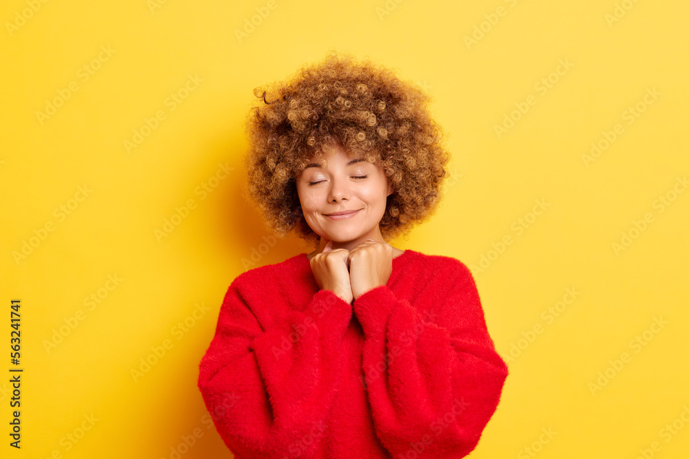 Indoor shot of cute curly haired young lady keeps clenched fists together under chin, dreams with closed eyes, wears casual red pullover, poses over yellow wall Indoor shot of cute curly haired young lady keeps clenched fists together under chin, dreams with closed eyes, wears casual red pullover, poses over yellow wall