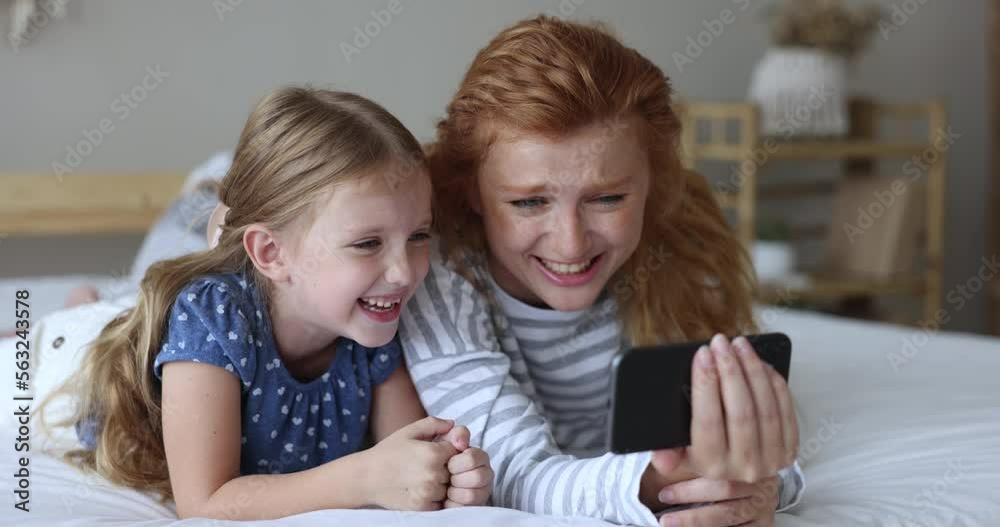 Beautiful little 6s girl and young redhead mother lying on bed staring at cellphone screen laughing while watching funny videos on internet, close up. Family pastime using modern wireless tech, fun Beautiful little 6s girl and young redhead mother lying on bed staring at cellphone screen laughing while watching funny videos on internet, close up. Family pastime using modern wireless tech, fun