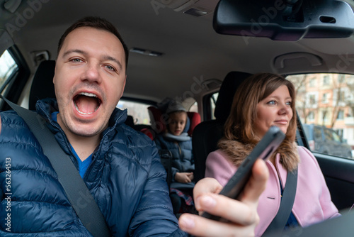 Woman is driving car with excited happy man sitting in passenger seat and looking at camera. Enjoying road trip together. Married couple with children on trip. Concept of travel, love and family