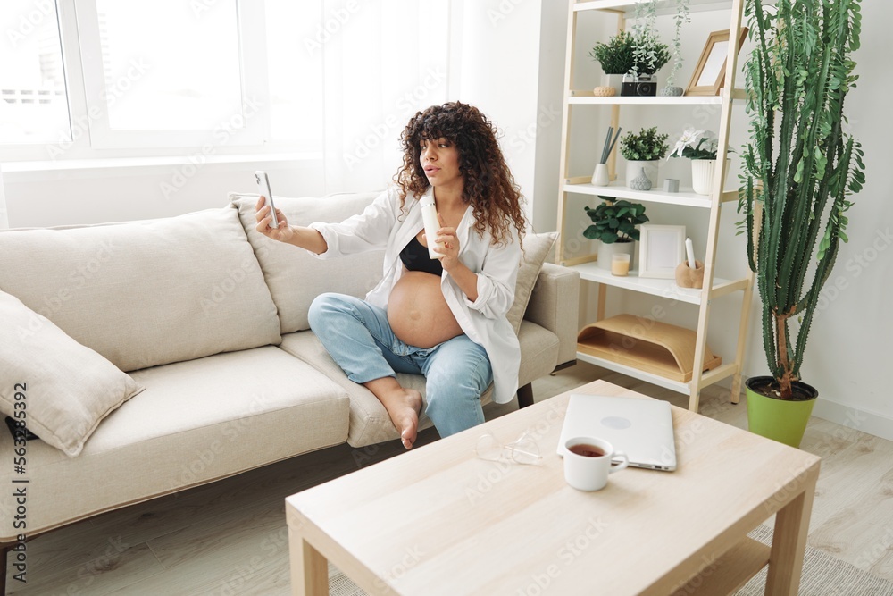 Pregnant woman smile blogger showing baby feeding bottle filming herself on phone, video chat sitting on sofa at home freelancer in last month of pregnancy Pregnant woman smile blogger showing baby feeding bottle filming herself on phone, video chat sitting on sofa at home freelancer in last month of pregnancy