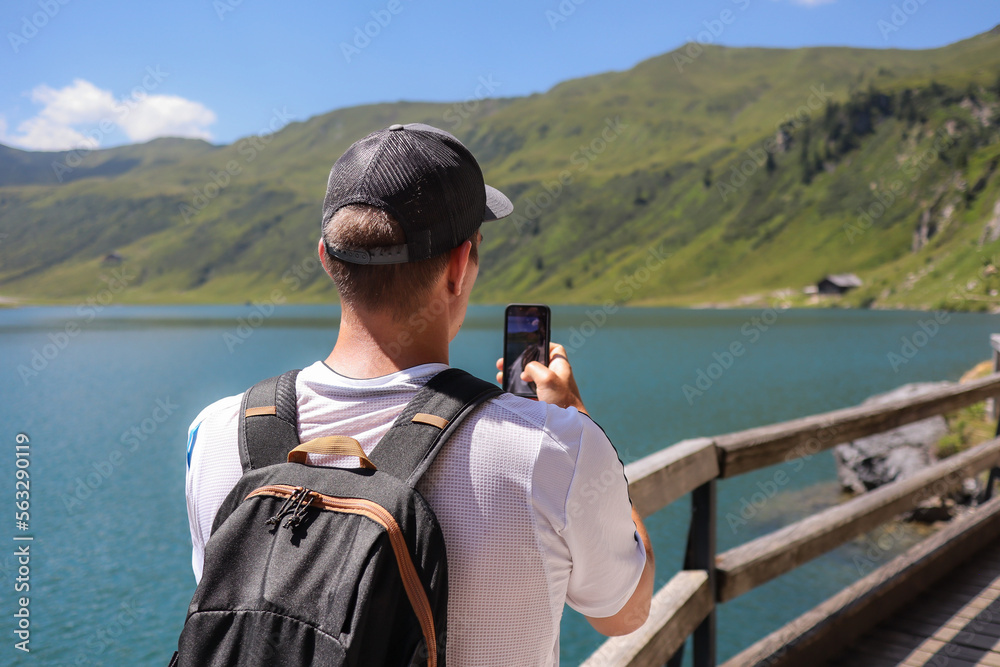 Obraz premium Caucasian Man from Behind with Smartphone in European Nature. Young Adult with Backpack and Cap Takes Picture with Phone of Scenic Outdoor Scene of Alpine Lake during Summer Sunny Day in Austria.