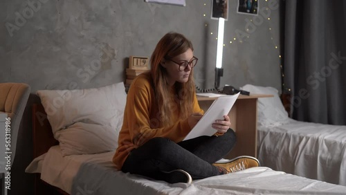 student in glasses sits on a bed in college dormitory holding sheets of paper in his hands enters information into a search engine on his smartphone. the concept of preparing for the exam. copy space. photo